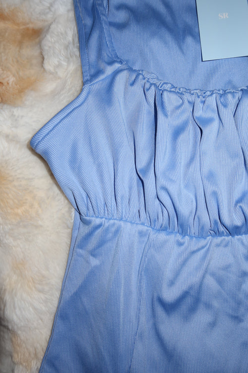 A close-up of a light blue ribbed fabric garment laid flat on a cream faux-fur surface, showing gathered ruching and a small light blue tag in the upper left corner.